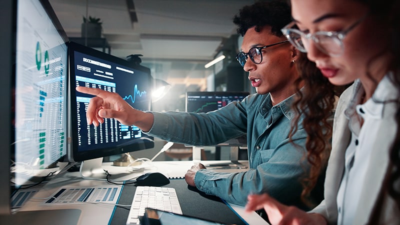 Two individuals in a modern office collaborate at computers displaying data. A young man points at his screen while a young woman focuses on a tablet.