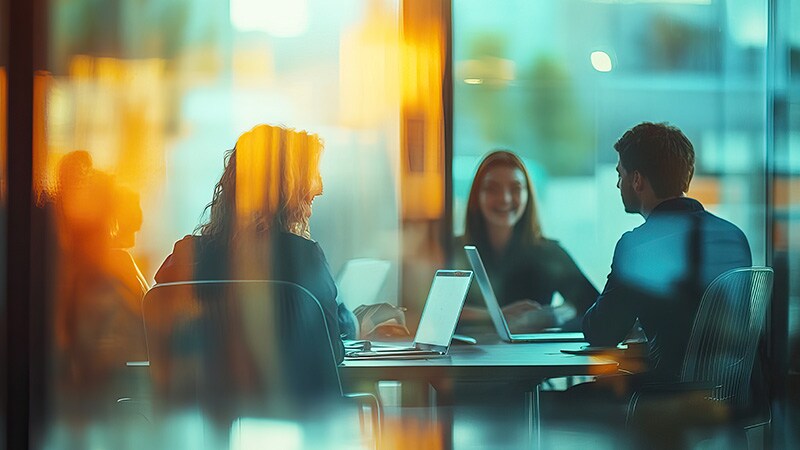 A group of three people engaged in a discussion around a conference table, with laptops and documents present, in a warm, well-lit office environment.