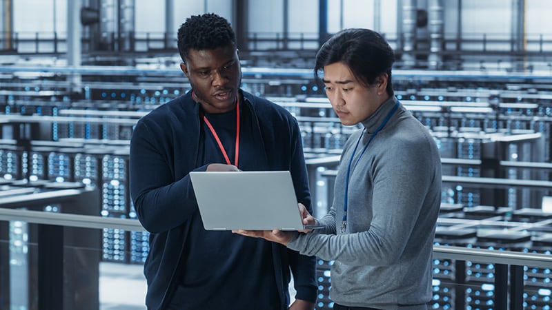 Two men discussing a laptop in a data center, surrounded by server racks with blue lights.