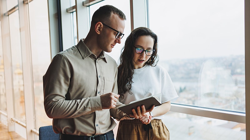 A man and a woman are discussing over an open notebook by a large window, with a cityscape visible in the background.