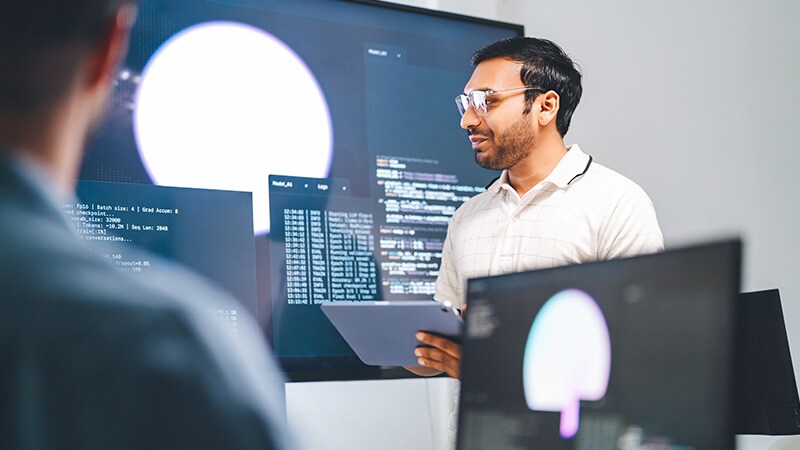 A female presenter showcases an AI Assistant graphic on a large screen in a modern office, while four attendees engage in the presentation.