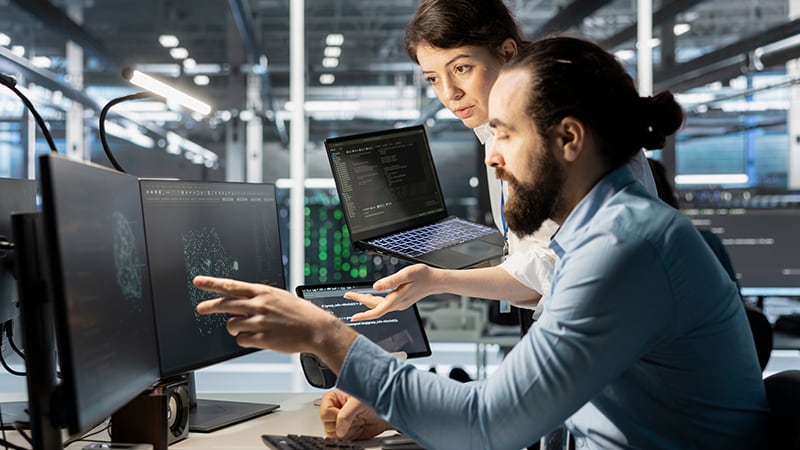Two colleagues collaborate in a tech office; one points at a computer screen while holding a laptop, with multiple monitors displaying data in the background.