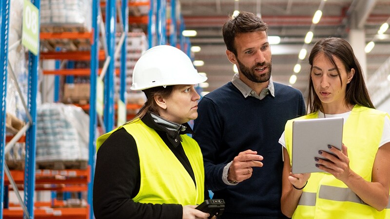Three individuals, two in safety vests examining a tablet together, engaged in a discussion or analysis.