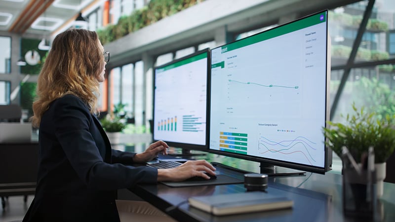 A woman with blonde hair sits at a modern desk, using two large monitors displaying data graphs in a bright office with green plants in the background.