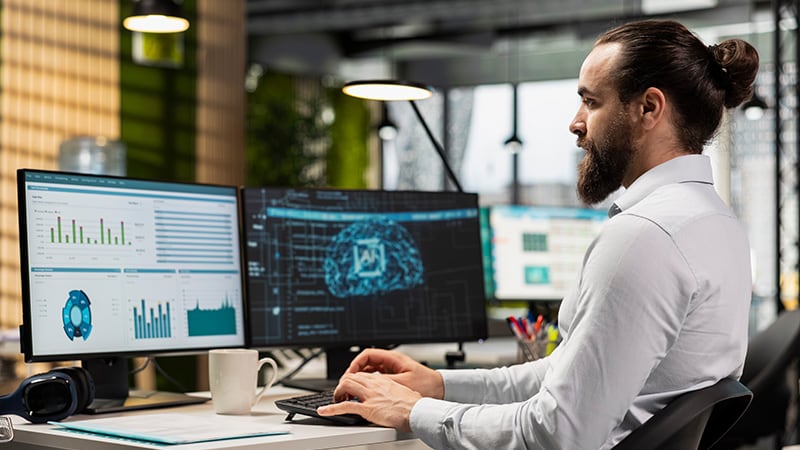 A man in a light shirt works at a desk with two monitors displaying data graphs and a digital brain graphic, with a mug and headphones nearby.